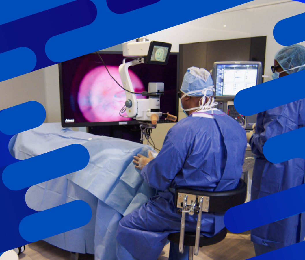 Surgeon performing vitreoretinal surgery using an operating microscope and digital imaging system, with large display screen showing a close-up of the eye