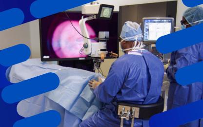 Surgeon performing vitreoretinal surgery using an operating microscope and digital imaging system, with large display screen showing a close-up of the eye